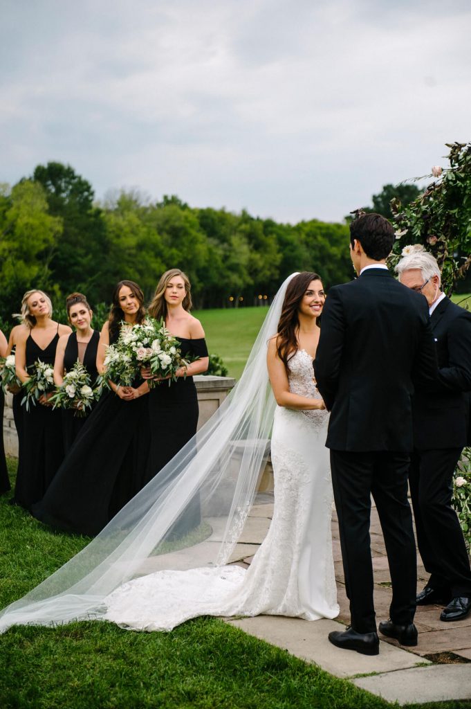 bride looks at groom with bridesmaids in black dresses in background great marsh estate