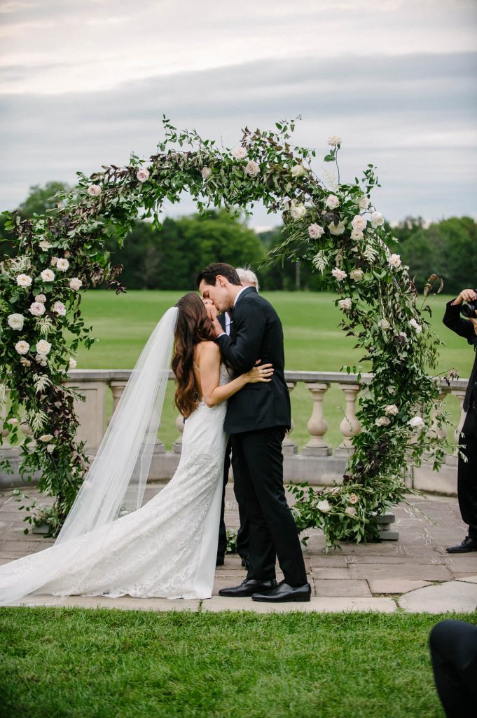 bride and groom kiss in front of circular flower arch great marsh estate