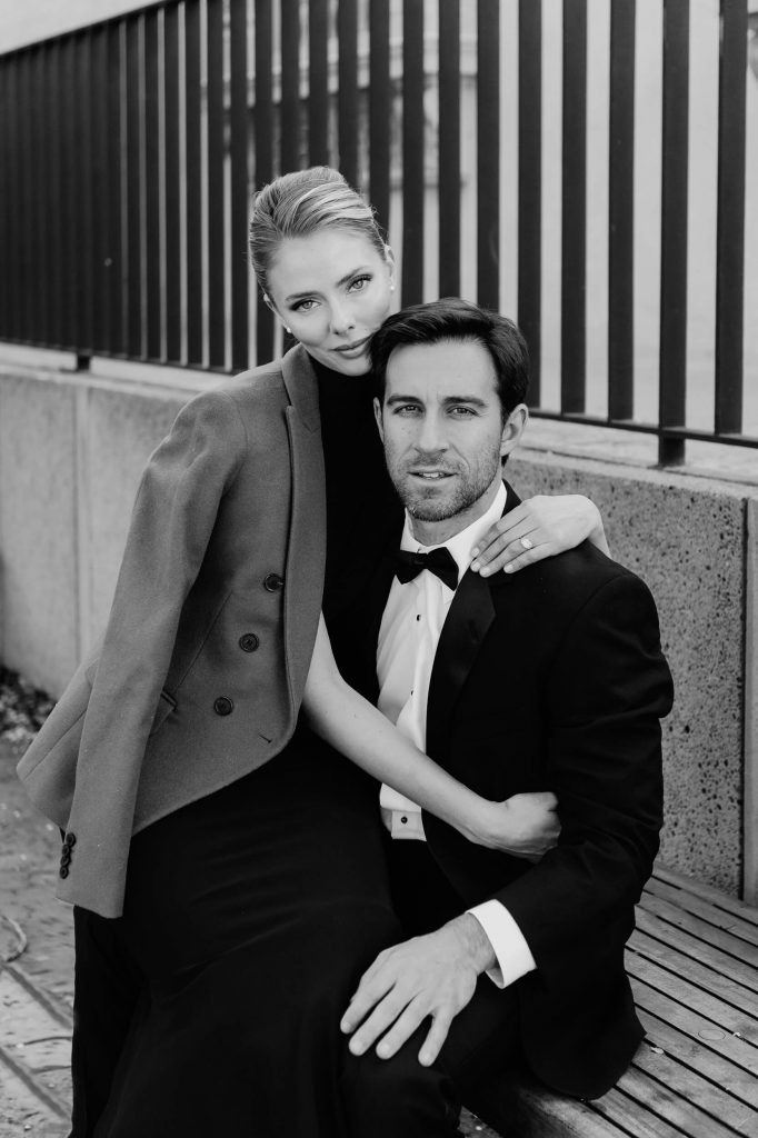 woman in peacoat and formal black gown embraces fiancee in tuxedo for black and white classic engagement photo