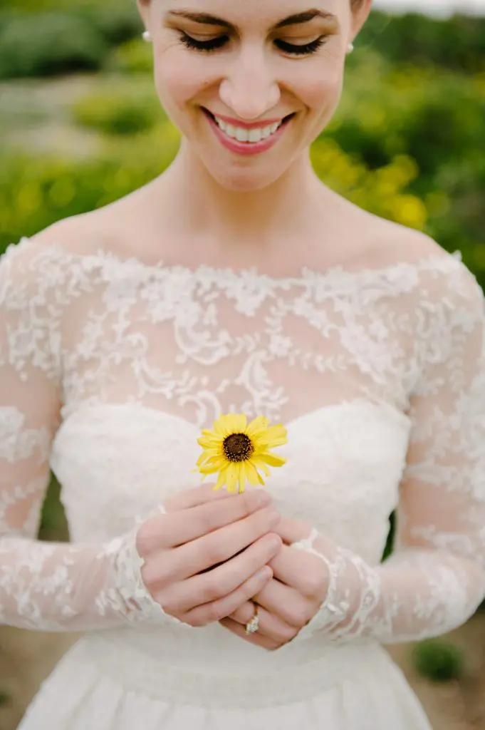 bride in gown with long train and lace sleeves holding yellow wildflower poses at scripps coastal reserve