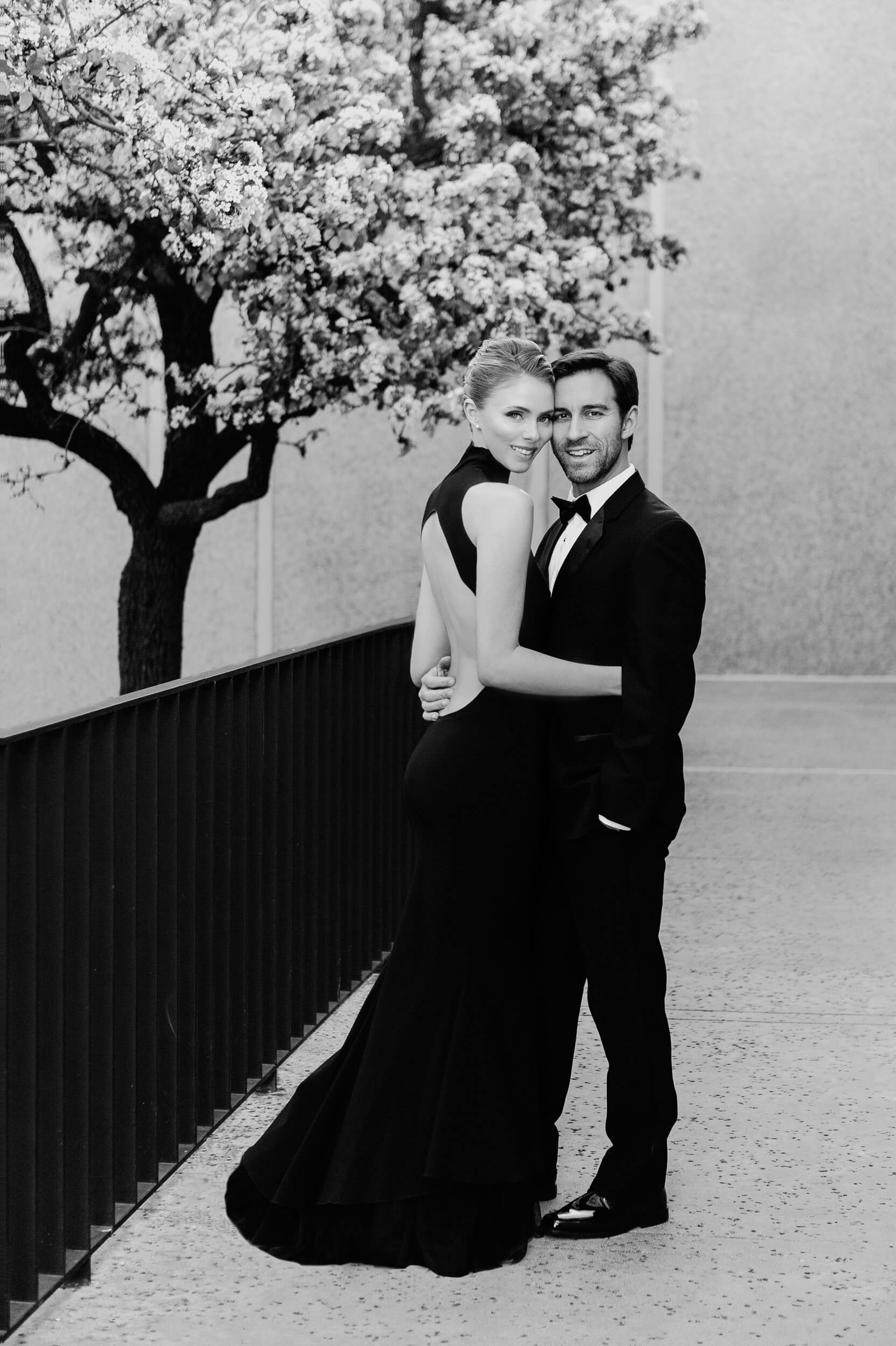 couple in formalwear pose near flowering tree and black railing balboa park classic engagement photo