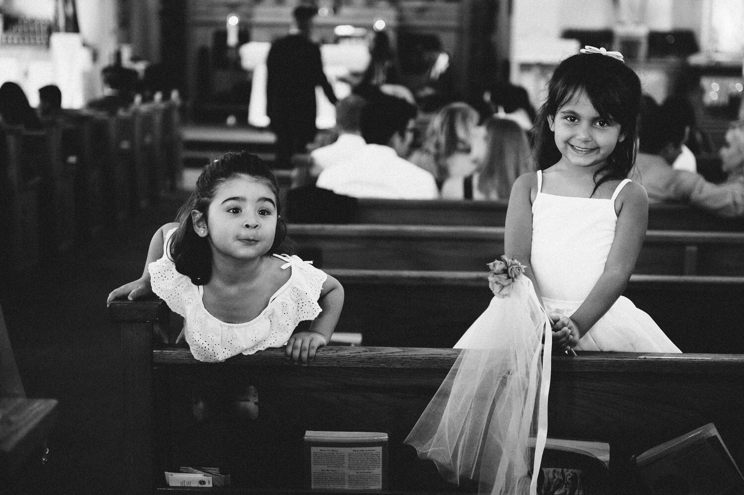 flower girls in white dresses stand in church pew and smile at camera