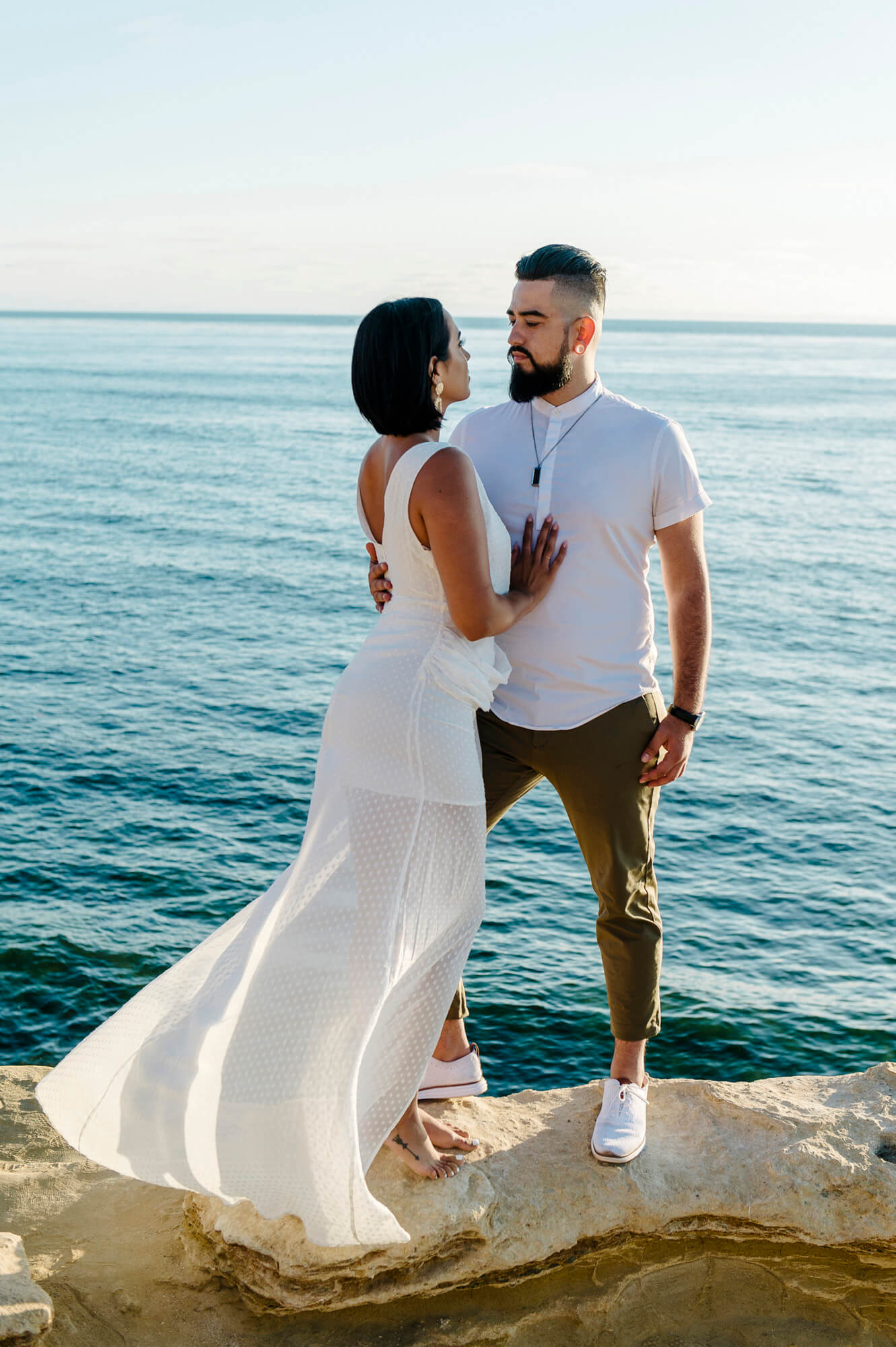 woman in long white beach dress embraces fiancee in t-shirt and khakis at sunset cliffs engagement photo