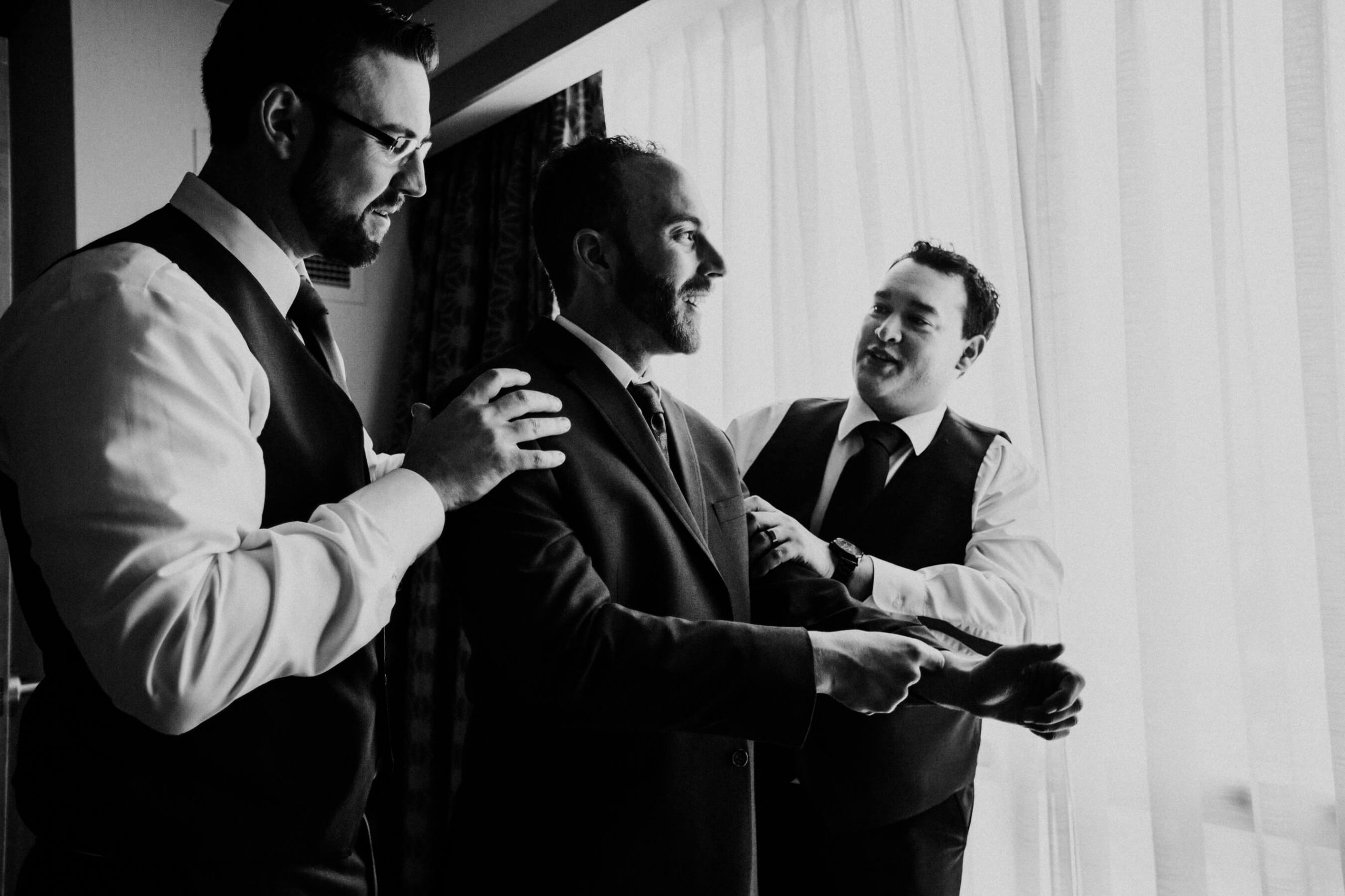 groomsmen help groom with suit jacket in hotel room