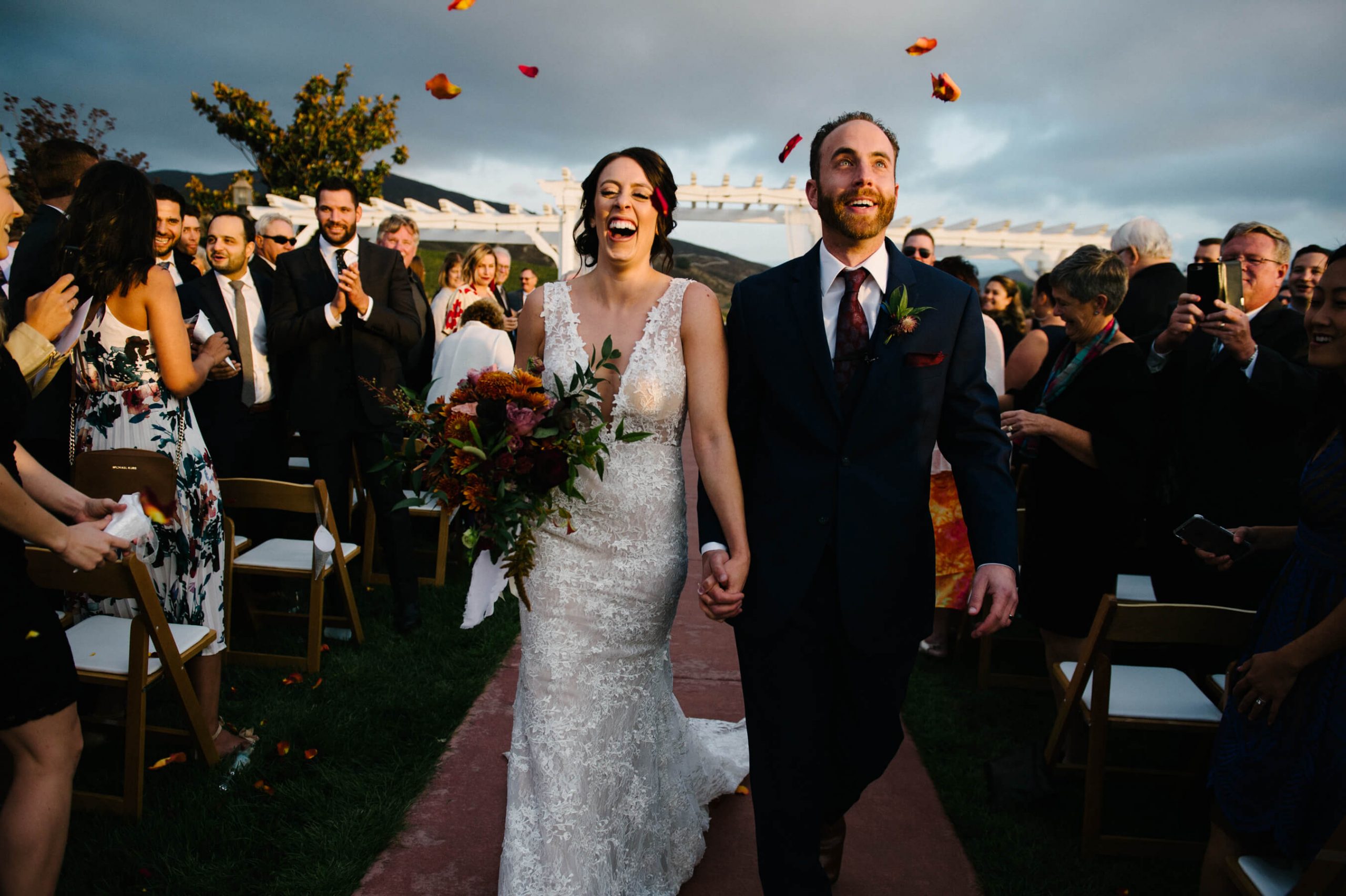 guests throw rose petals in air as bride and groom process down aisle leoness cellars