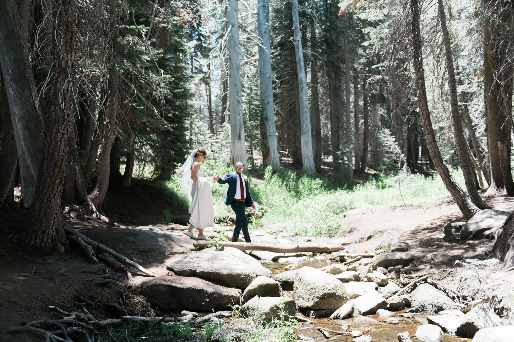 groom holding bride's hand as she walks over creek during yosemite adventure elopement
