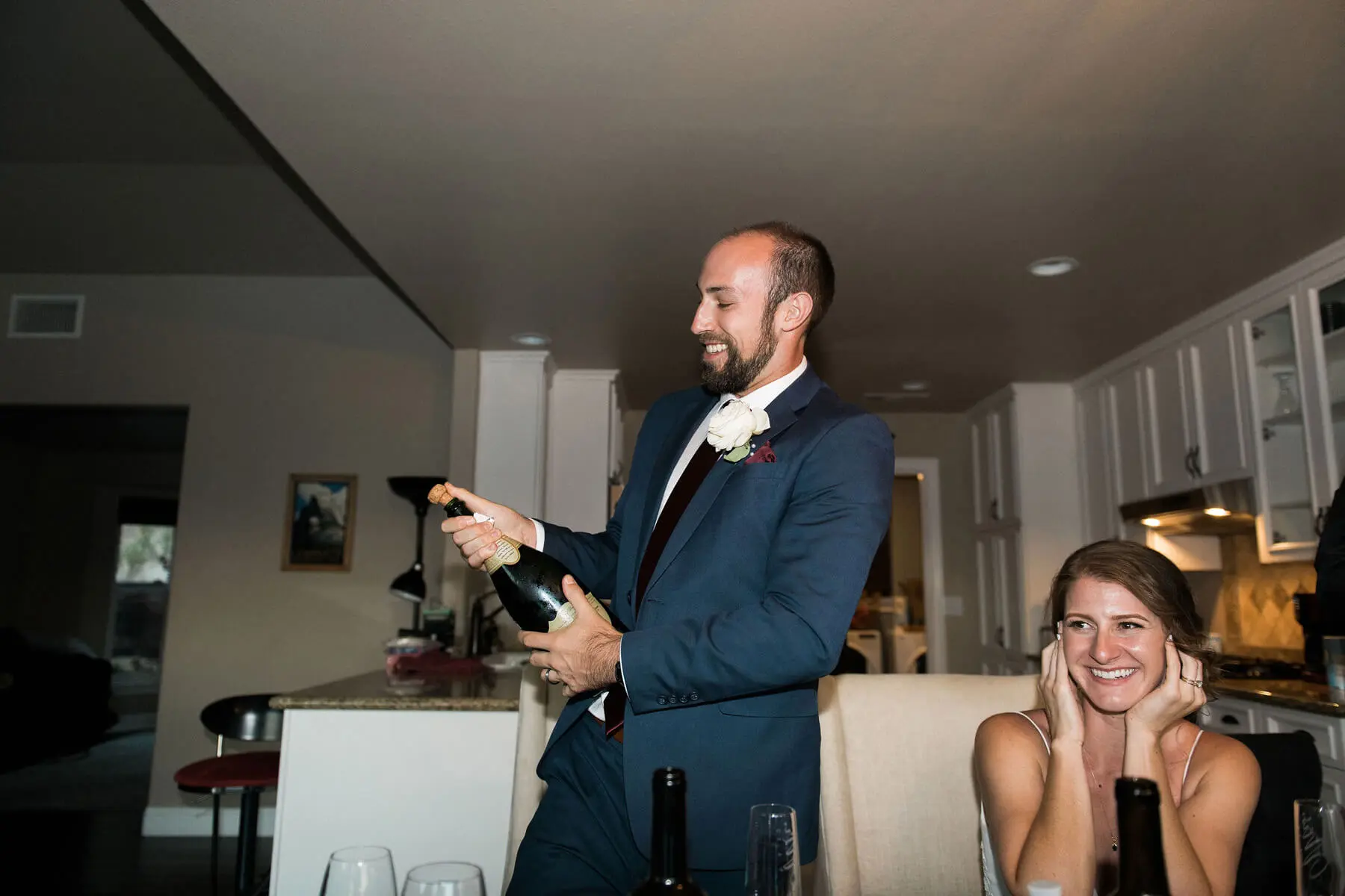 groom popping bottle of champagne while bride smiles and covers ears